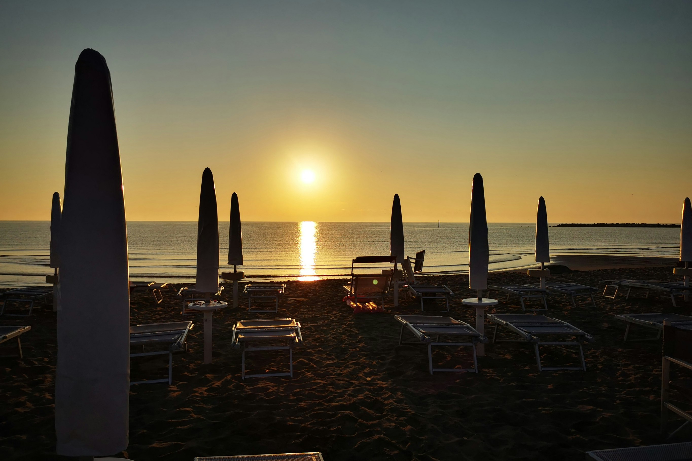 Sonnenuntergang am Strand: Liegestühle und Sonnenschirme auf dem Sand, im Hintergrund das Meer mit tiefstehender Sonne.