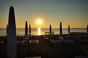 Sonnenuntergang am Strand: Liegestühle und Sonnenschirme auf dem Sand, im Hintergrund das Meer mit tiefstehender Sonne.