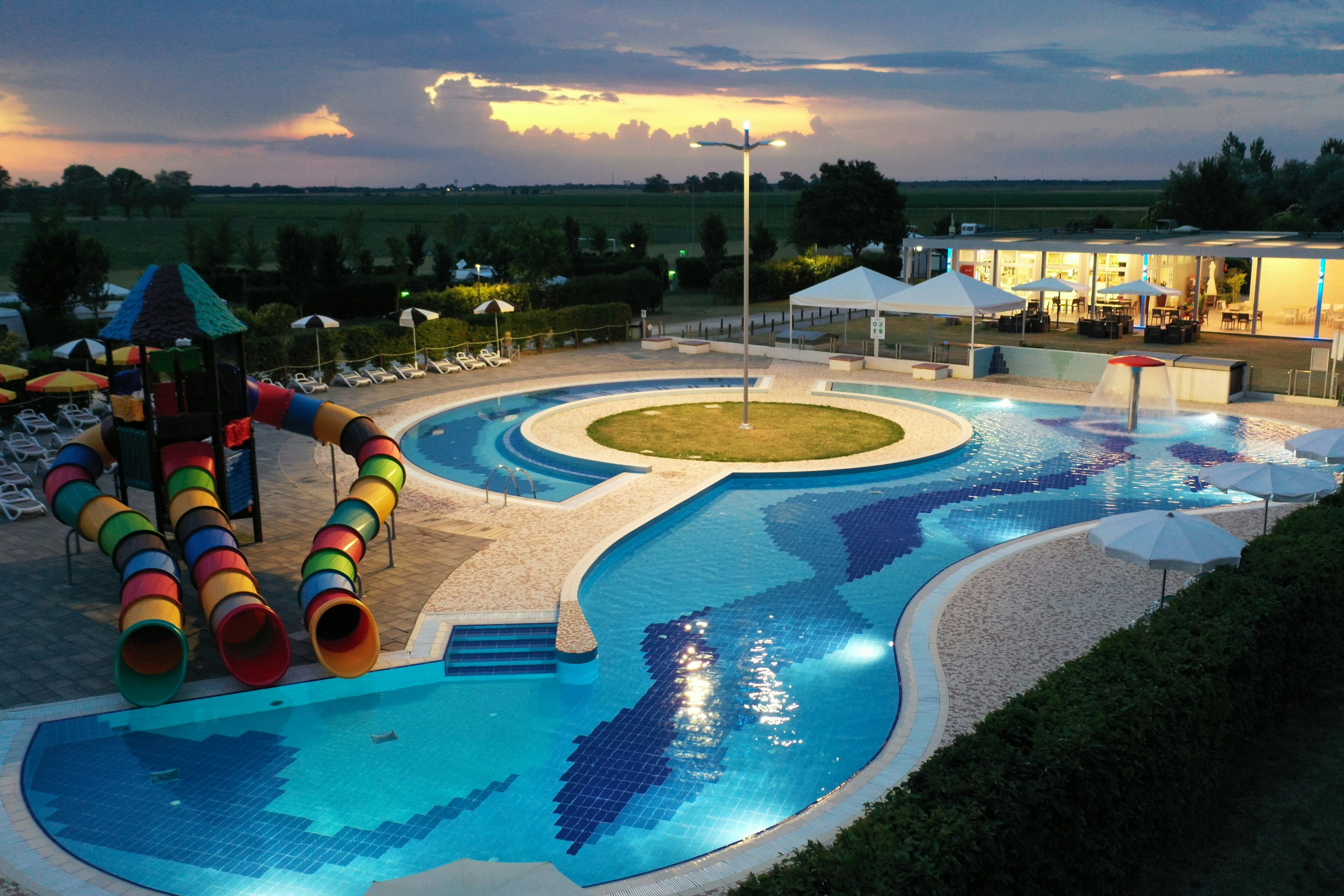 Großer Campingpool mit Rundbecken, Liegen und bunten Wasserrutschen, im Hintergrund Zelte und Gebäude bei Sonnenuntergang.