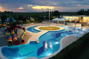 Großer Campingpool mit Rundbecken, Liegen und bunten Wasserrutschen, im Hintergrund Zelte und Gebäude bei Sonnenuntergang.