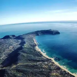 Luftaufnahme einer Küste mit Bucht und Sandstrand, umgeben von Felsen und Meer unter klarem Himmel.