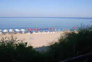 Blick auf einen Strand am See mit Liegen, Sonnenschirmen und Sand, im Hintergrund ruhiges Wasser.