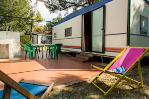 Wohnwagen mit Terrasse, grünen Gartenstühlen und Liege, daneben ein kleiner Pool und Bäume im Hintergrund.