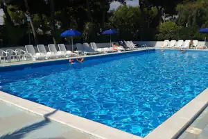 Blauer Camping-Pool mit Liegen, Sonnenschirmen und Bäumen im Hintergrund, im Becken schwimmt eine Person.