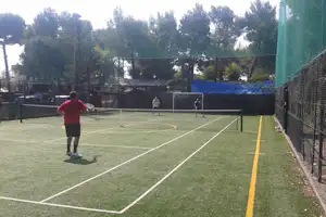 Tennisplatz auf dem Campinggelände mit Netz, grüner Kunstrasenfläche und Umzäunung, im Hintergrund Bäume und weitere Plätze.