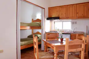 Blick in den Wohnbereich mit Esstisch, Holzstühlen und Küche; dahinter ein Schlafbereich mit Etagenbett und Fenster.
