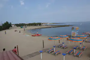 Sandstrand mit bunten Sonnenschirmen und Liegen, daneben ein Boot im Wasser und ein Uferweg mit Bäumen.