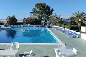 Großer Camping-Pool mit weißen Liegen und Sonnenschirmen, im Hintergrund Gebäude, Bäume und Palmen im Außenbereich.