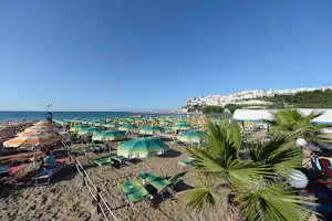 Reihe von Strandliegen und Sonnenschirmen am Sandstrand, im Hintergrund das Meer und ein Küstenabschnitt mit weißen Häusern.