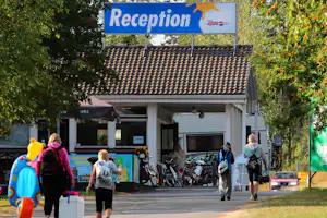 Rezeption eines Campingplatzes mit blauem Schild, Weg und Bäumen; im Hintergrund Fahrräder und ein Gebäude.