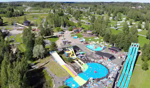 Luftaufnahme eines Campingplatzes mit mehreren Schwimmbecken, Wasserrutschen, Liegen und Wegen zwischen grünen Wiesen und Bäumen.