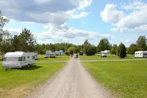 Wohnwagen auf einem Campingplatz, mittig ein Schotterweg durch die grüne Wiese, Bäume und weitere Fahrzeuge am Rand.