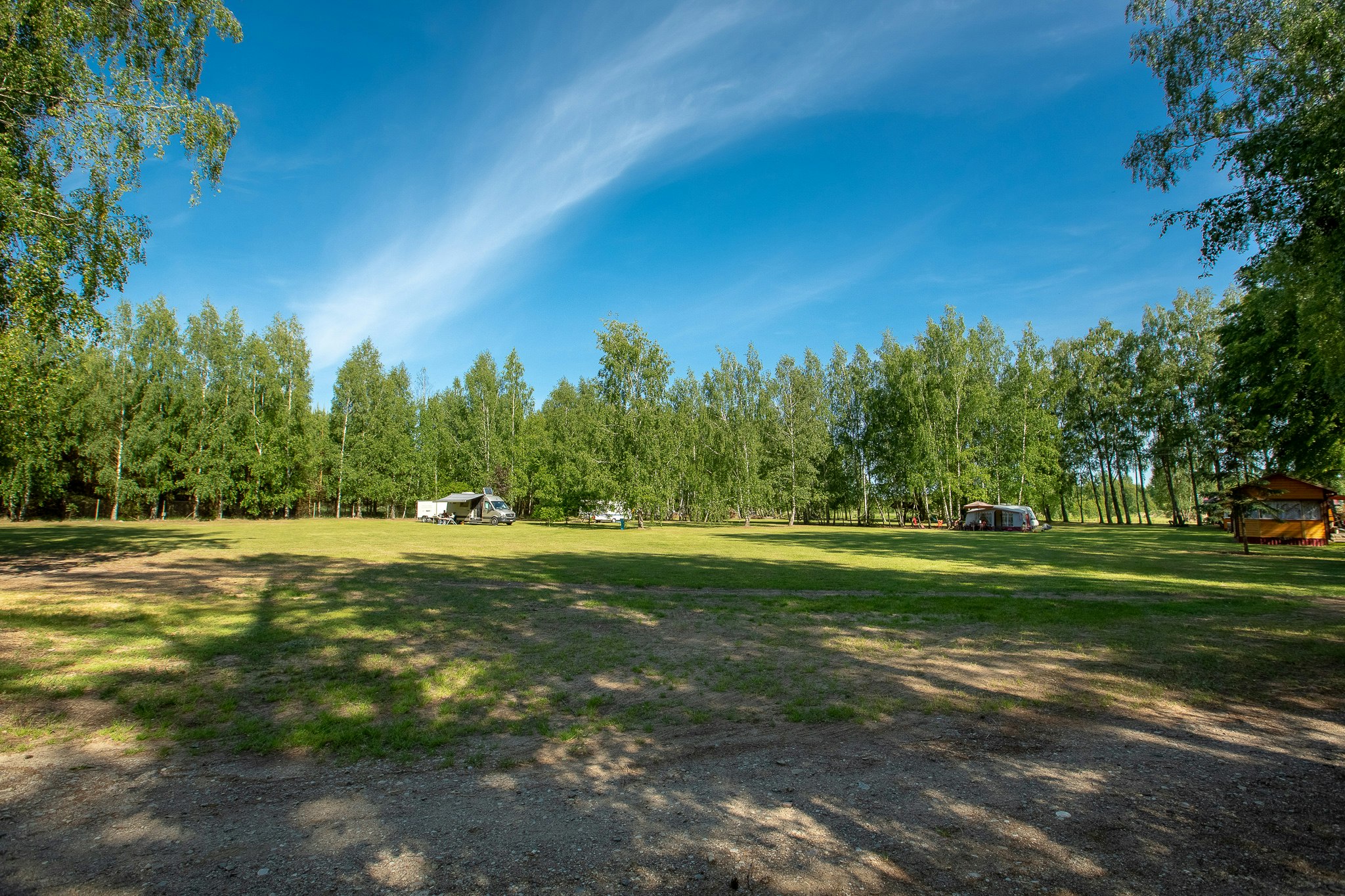 Große Wiese auf dem Campingplatz mit mehreren Wohnwagen und Zelten, umgeben von hohen Bäumen und blauem Himmel.