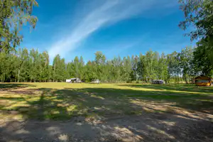 Große Wiese auf dem Campingplatz mit mehreren Wohnwagen und Zelten, umgeben von hohen Bäumen und blauem Himmel.