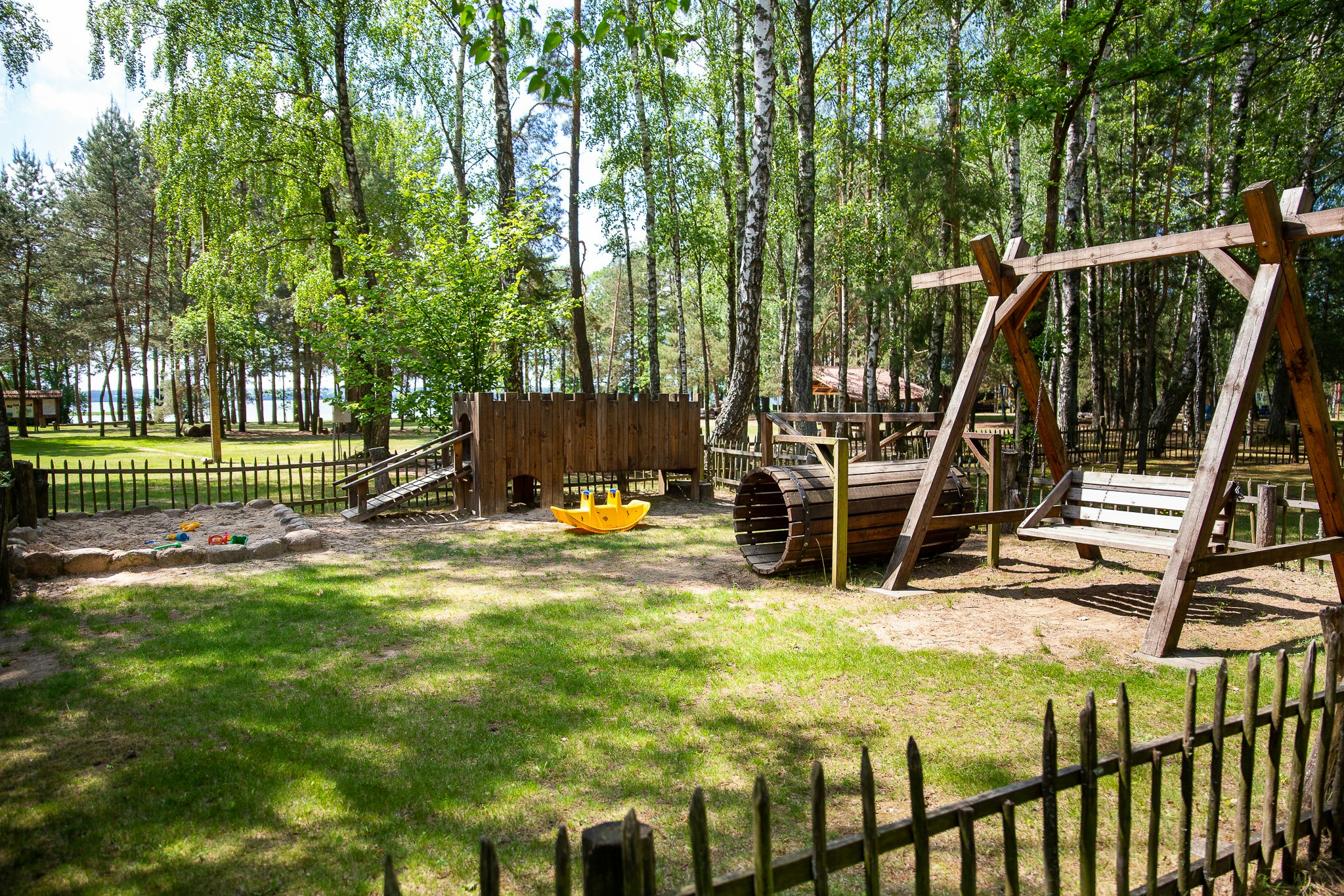 Spielplatz am Campingplatz mit Holzschaukel, Sandfläche und Sitzbank, umgeben von Bäumen und einem eingezäunten Bereich.