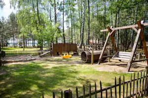 Spielplatz am Campingplatz mit Holzschaukel, Sandfläche und Sitzbank, umgeben von Bäumen und einem eingezäunten Bereich.