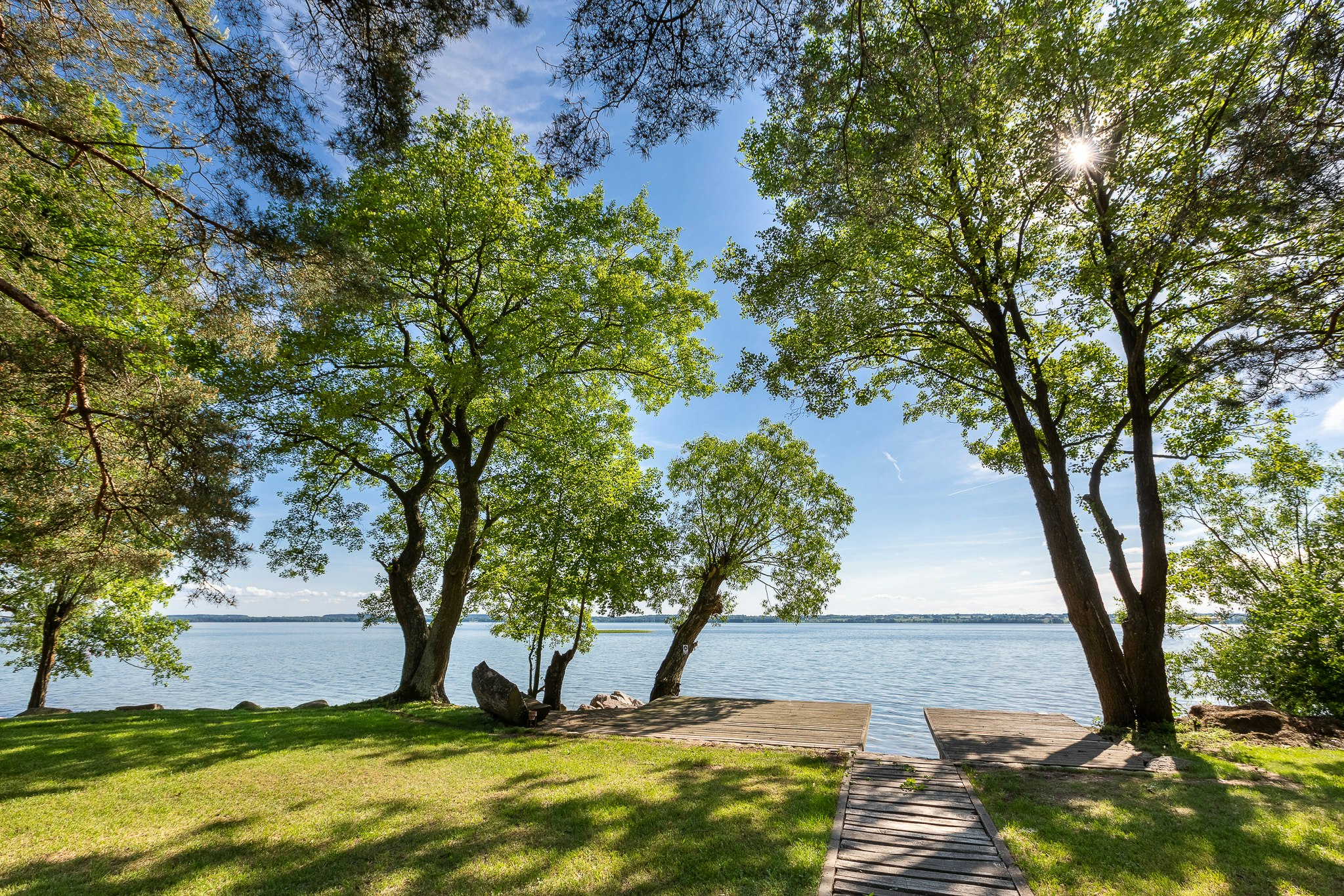 Uferpromenade am See mit Holzsteg, Rasenfläche und Bäumen im Vordergrund, Blick über das Wasser.