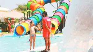 Kinder und Erwachsene am Pool mit bunten Wasserrutschen, im Hintergrund Palmen und ein weiteres Becken.