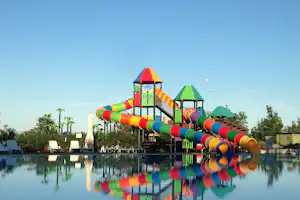 Bunte Wasserrutschen am Pool eines Campingplatzes, mit Spielturm und Spiegelung im Wasser vor blauem Himmel.