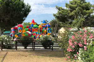 Buntes Hüpfburg-Spielgerät auf einem Campingplatz, umgeben von Bäumen, Blumenbeeten und einem Holzgeländer.