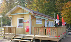 Gelbes Campinggebäude mit Rezeption, Holzterrasse und Markisen, im Hintergrund Bäume im Herbst.
