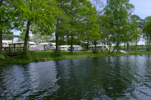 Campingplatz am See mit Wohnwagen und Zelten zwischen grünen Bäumen, im Vordergrund Wasser und Rasen.