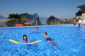 Schwimmbecken im Campingbereich mit gelben Schwimmnudeln, Kinder im Wasser und Blick auf Berge und Seeufer.