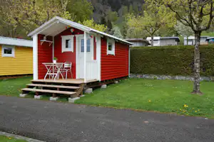 Rotes Mobilheim mit weißem Eingang und Terrasse, Stühle und Tisch vor dem Haus, umgeben von Rasen und Bäumen.