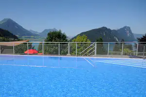 Blauer Swimmingpool mit Geländer und Blick auf grüne Berge und ein Tal, im Hintergrund Bäume und Wasserfläche.