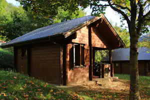 Holzhütte mit Satteldach und Eingangstür, davor Terrasse mit Stühlen, umgeben von Bäumen und Rasen im Campingbereich.