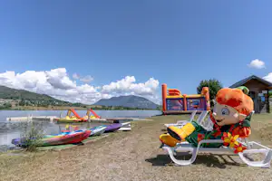 Kinderfahrzeug auf dem Campingplatz am See, daneben Schlauchboote und Ruderboote am Ufer, im Hintergrund Berge.