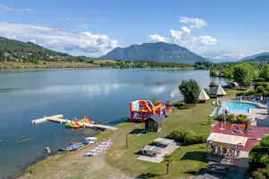 Blick auf den See mit Liegewiese, Sonnenschirmen und Pool am Campingplatz, im Hintergrund Berge und bewaldete Hänge.