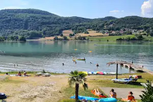 Seeufer mit Campingplatz: Badende im Wasser, Liegen und Sonnenschirme am Strand, Ruderboote auf dem ruhigen See.