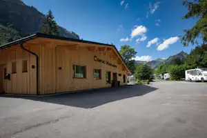 Holzgebäude auf dem Campingplatz mit Beschilderung, Asphaltweg und geparktem Wohnwagen vor Bergen und Bäumen.