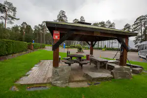 Holzüberdachter Grillplatz mit Steintisch und Bänken auf dem Campingplatz, Rasenfläche, Bäume und Wohnwagen im Hintergrund.