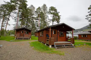 Holzhütten auf einem Campingplatz mit Rasenfläche, Kiesweg und Bäumen im Hintergrund.