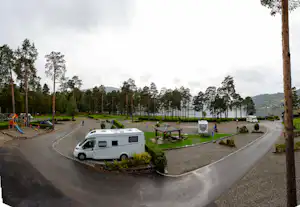 Weißes Wohnmobil auf einem Stellplatz im Campingplatz, mit Tisch und Bänken, Wegen und vielen Kiefern im Hintergrund.