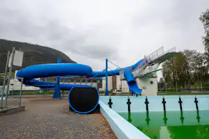 Wasserrutsche und Schwimmbecken auf dem Campingplatz, mit blauen Leitern, Zaun und Spielbereich am Rand.