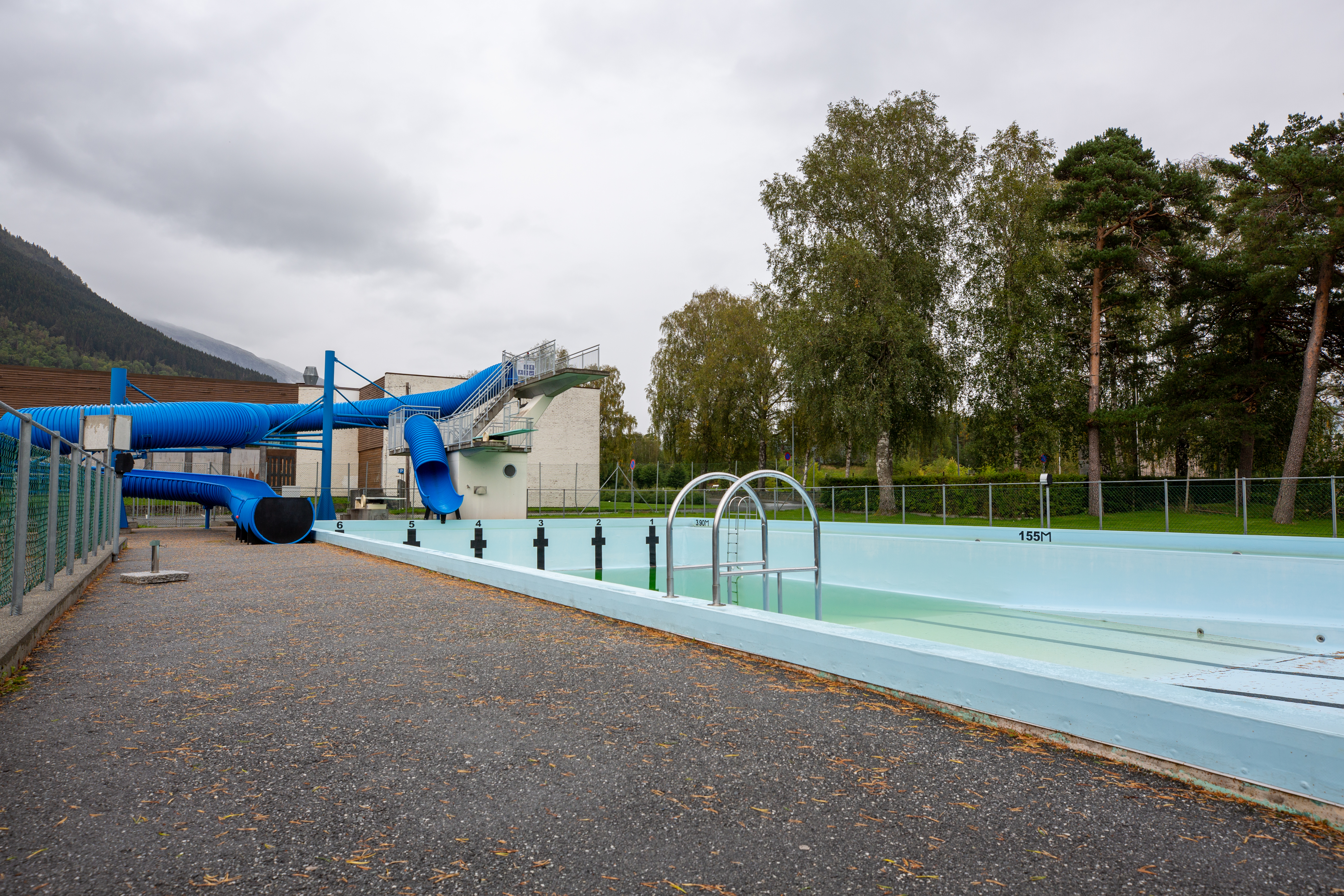 Schwimmbecken am Campingplatz mit Einfassung, Zaun und Wegen, im Hintergrund Bäume und Berge.