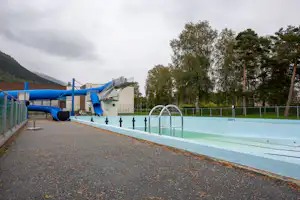 Schwimmbecken am Campingplatz mit Einfassung, Zaun und Wegen, im Hintergrund Bäume und Berge.