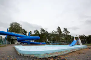 Blaues Wasserrutschen-Becken auf einem Campingplatz mit Wasserbahn, Umrandung und Zaun, im Hintergrund Bäume und Gebäude.