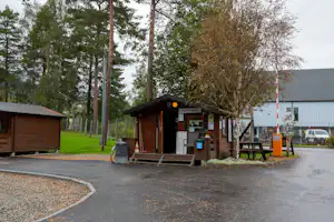 Holz-Rezeption am Campingplatz mit Schildern, Bank und Parkplatz, umgeben von Bäumen und einem kleinen Weg.