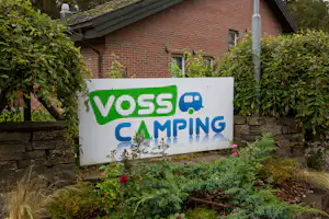 VOSS Camping Schild vor einem Backsteinhaus, umgeben von Hecken und Pflanzen im Gartenbereich.