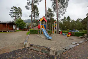 Spielplatz am Campingplatz mit Rutsche, Sandfläche und Wegen, im Hintergrund Bungalows und Bäume.