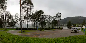 Campingplatz mit gepflastertem Weg, Wohnwagen und Wohnmobil am Rand, umgeben von hohen Bäumen und Hügeln im Hintergrund.