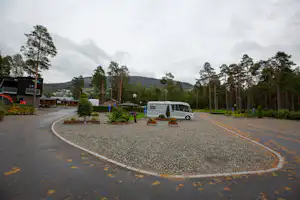 Einfahrt eines Campingplatzes mit Wohnmobil auf dem Parkplatz, geschwungene Straße, Bäume und Hecken entlang der Anlage.