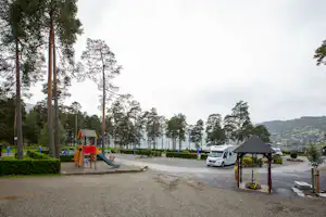 Campingplatzweg zwischen hohen Kiefern, daneben ein Spielbereich und geparkte Autos, im Hintergrund Hügel.