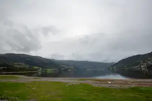 Blick auf einen See im Tal, grünes Ufer und Wiese am Campingplatz, umgeben von Hügeln und bewölktem Himmel.