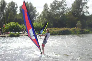 Windsurfer auf dem See mit rotem und blauem Segel, im Hintergrund Bäume und weitere Personen am Ufer.