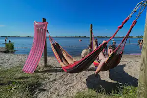 Bunte Hängematten am Sandstrand am See, daneben ein aufblasbarer Schwimmreifen und weitere Gäste im Hintergrund.